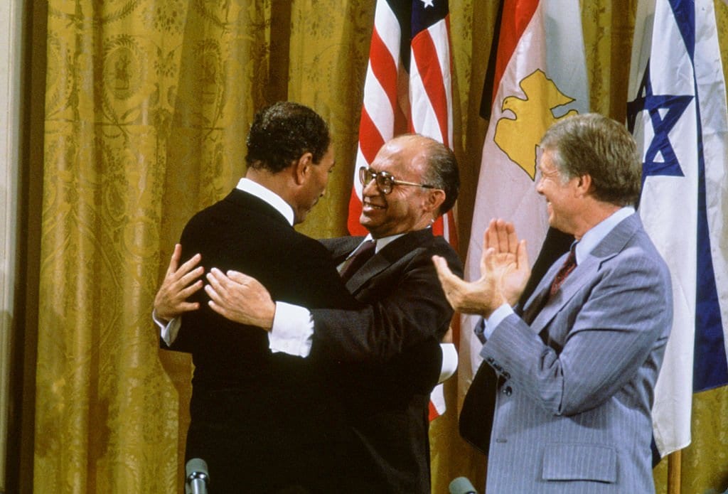 O presidente egípcio Anwar al-Sadat e o primeiro-ministro israelense Menachem Begin se abraçam, enquanto o presidente americano Jimmy Carter aplaude depois de assinarem os acordos de Camp David na Sala Leste da Casa Branca, 18 de setembro de 1978, em Washington, DC. [David Hume Kennerly / Getty Images]