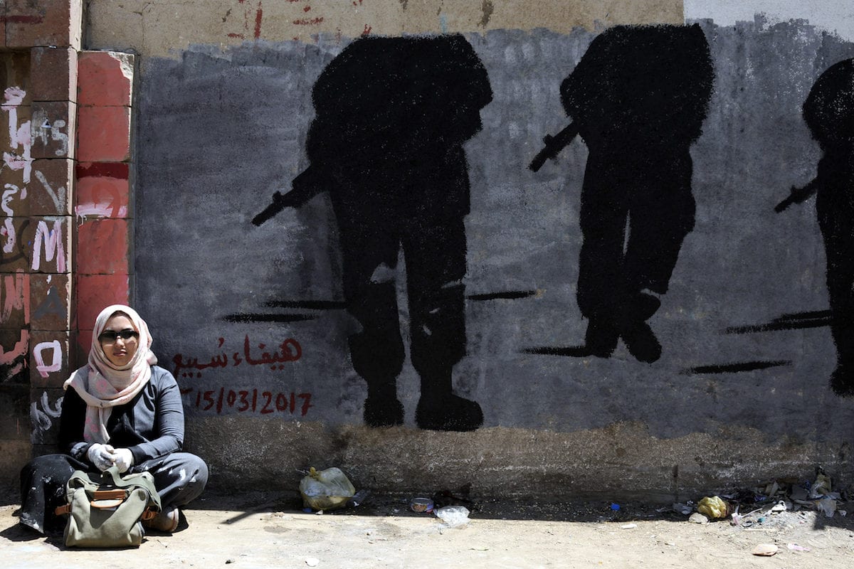 Mulher sentada em frente a uma parede com pinturas de silhuetas segurando rifles em Sanaa, Iêmen, em 15 de março de 2017. [Agência Mohammed Hamoud/Anadolu]