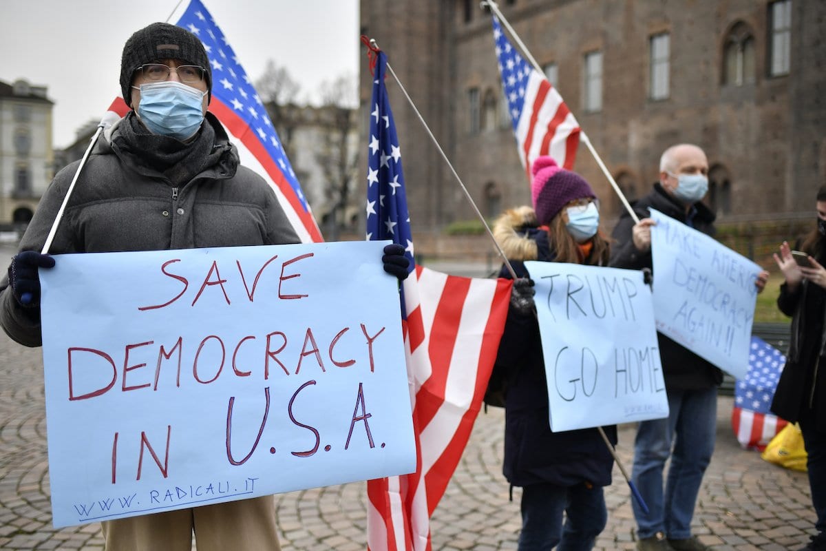 Pessoas usando máscaras protetoras acenando bandeiras americanas e segurando cartazes de protesto onde se lê “Trump, vá para casa” e “Salve a democracia nos EUA”, na Piazza Castello, em 09 de janeiro de 2021, em Torino, Itália. [Stefano Guidi/Getty Images]