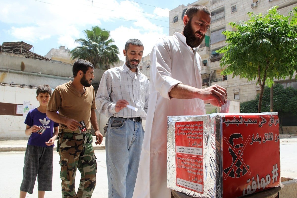 Homens sírios fingem que estão votando durante uma eleição simulada que pede que o "criminoso" presidente sírio Bashar al-Assad seja destituído de sua nacionalidade síria, em 3 de junho de 2014 na maioria cidade controlada pelos rebeldes de Aleppo. [BARAA Al-Halabi/ AFP via Getty Images]
