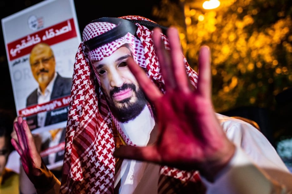 Um manifestante usa uma máscara representando o príncipe herdeiro saudita, Mohammad Bin Salman, com as mãos pintadas de vermelho ao lado de pessoas segurando pôsteres do jornalista saudita Jamal Khashogg, em 25 de outubro de 2018. [Yasin Akgum/AFP via Getty Images]
