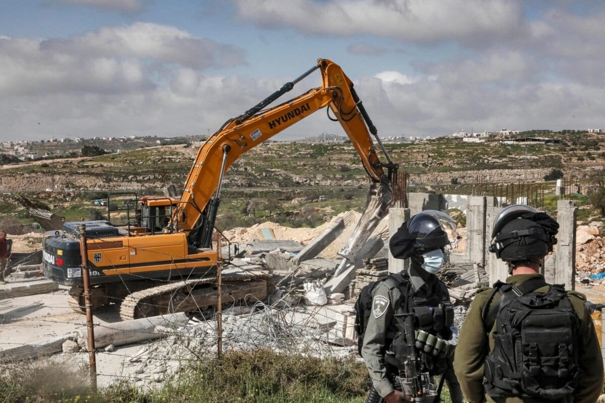 Um soldado israelense e um guarda de fronteira aguardam enquanto uma escavadeira destrói uma casa palestina localizada na área C a sudeste de Hebron, na Cisjordânia ocupada, em 8 de março de 2021. [Hazem Bader/AFP via Getty Images]