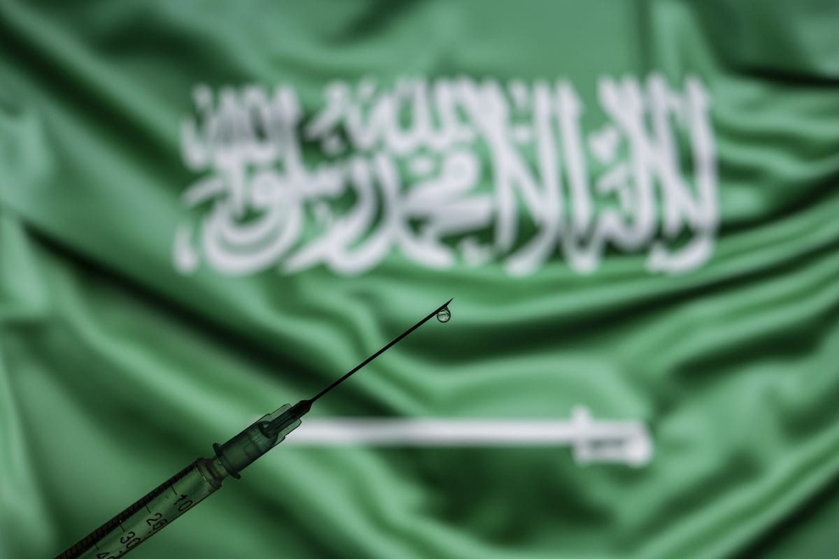 Uma seringa de vacina com uma gota na ponta da agulha é vista na frente de uma tela de computador que mostra a bandeira nacional da Arábia Saudita. [Ali Balıkçı - Agência Anadolu]