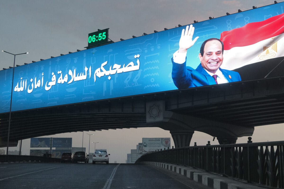 Carros passam ao longo de um viaduto sob um outdoor eletrônico gigante que mostra um banner representando o presidente Abdel Fattah al-Sisi ao lado de uma mensagem desejando viagens seguras, no Cairo, em 15 de janeiro de 2021 [Amir Makar/AFP via Getty Images]