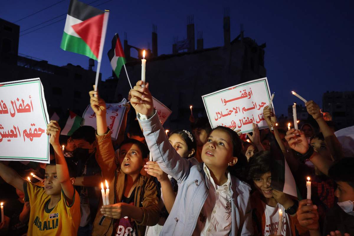 Crianças palestinas seguram velas durante um comício em meio às ruínas de casas destruídas pelos ataques israelenses, na Cidade de Gaza, em 24 de maio de 2021. Slogans em árabe dizem: “Pare os crimes de ocupação da Palestina [Mohammed Abed/AFP via Getty Images]
