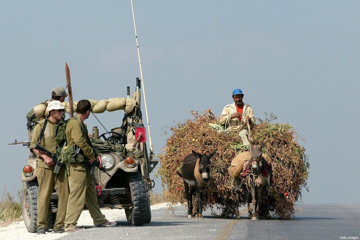 Um homem palestino usa uma carroça de burro para transportar forragem animal para seu rebanho enquanto passa por soldados israelenses que guardam a estrada, em 6 de agosto de 2002, ao redor da cidade de Qalqilya, na Cisjordânia [David Silverman/Getty Images]

