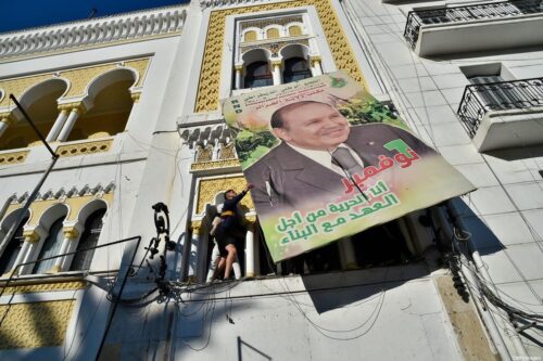 Manifestantes argelinos derrubam um grande outdoor com uma foto de seu atual presidente Abdelaziz Bouteflika, durante uma manifestação contra sua candidatura para um quinto mandato, em 22 de fevereiro de 2019 em Argel [Ryad Kramdi/AFP via Getty Images]