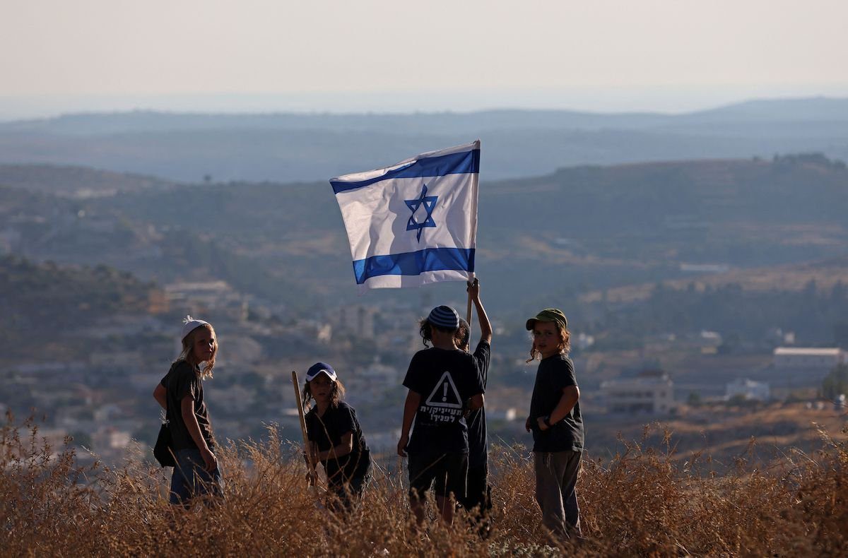Filhos de colonos israelenses agitam uma bandeira nacional perto do assentamento de Bat Ayin na Cisjordânia ocupada, em 21 de junho de 2021, enquanto ativistas de direita e líderes colonos realizam 14 marchas simultâneas para impedir o "controle árabe" na Área C. [Emmanuel Dunand/AFP via Getty Images]
