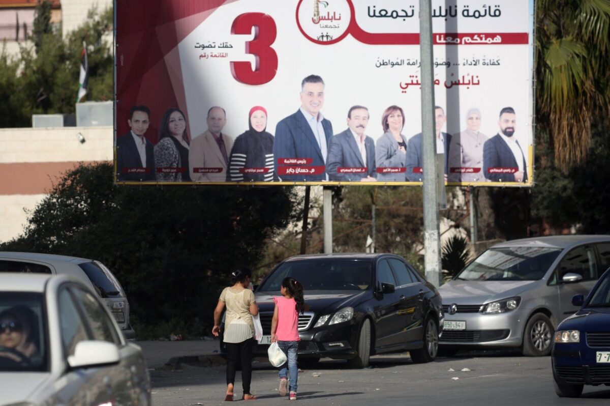 Duas meninas descem uma rua passando por um cartaz de campanha exibindo candidatos antes das eleições municipais na cidade de Nablus, no norte da Cisjordânia, em 10 de maio de 2017 [JAAFAR ASHTIYEH/AFP via Getty Images]