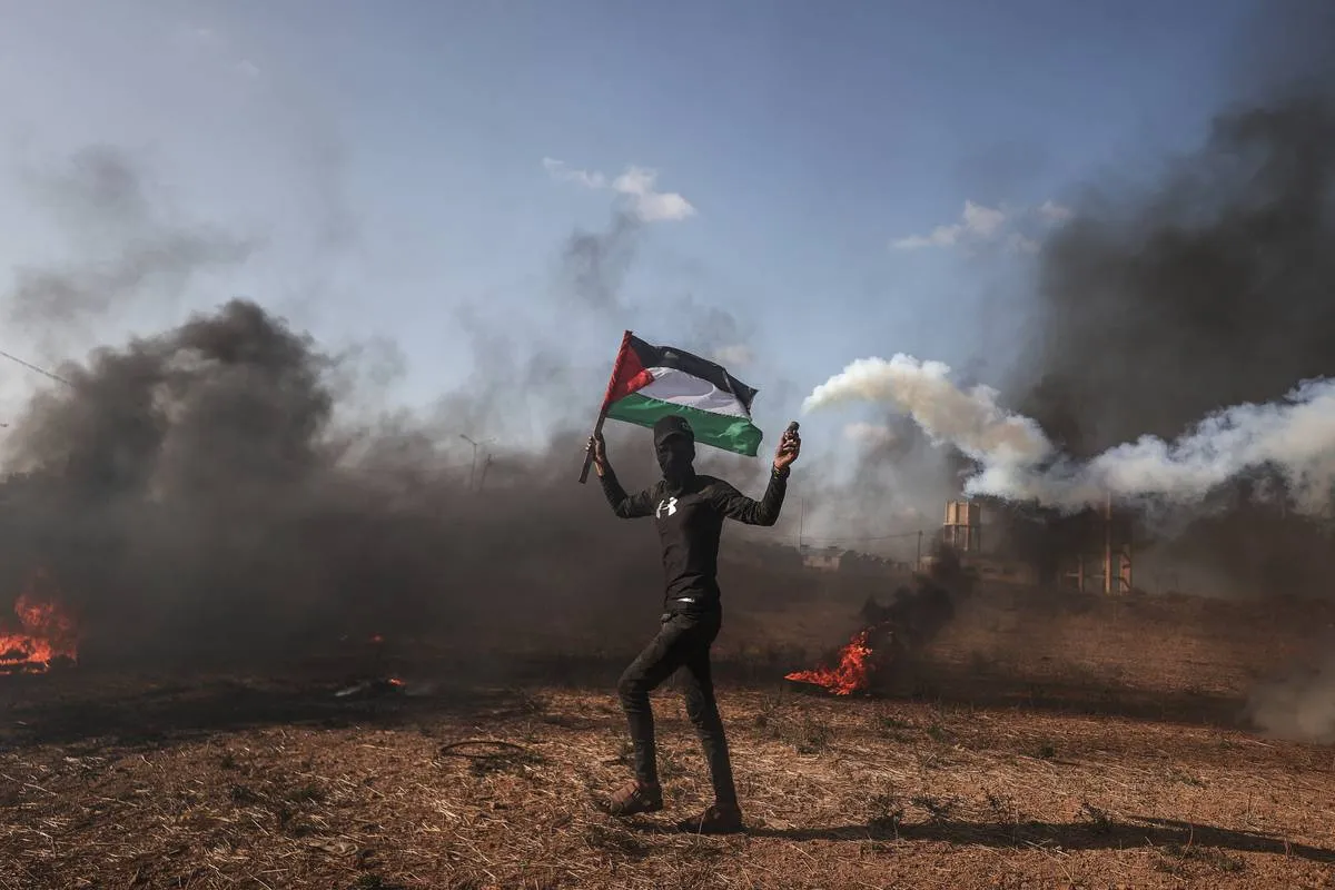 Homem de Gaza segura a bandeira da Palestina. Pneu é incendiado em protesto  contra a morte de palestinos pelas forças israelenses, na Faixa de Gaza em 19 de junho de 2023 [Ali Jadallah/Agência Anadolu]
