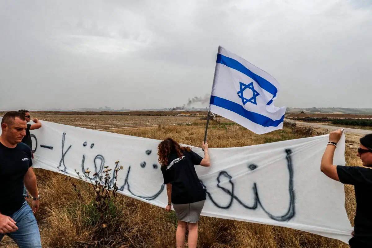 Foto tirada em 4 de maio de 2018 do kibutz de Nahal Oz, no sul de Israel, do outro lado da fronteira com a Faixa de Gaza, mostra colonos israelenses agitando uma bandeira israelense e segurando um banner [Ahmad Gharabli/AFP via Getty Images]
