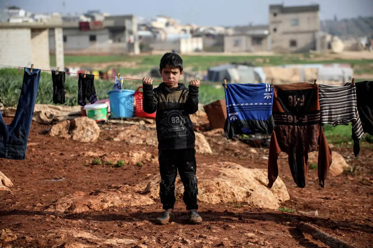 Uma criança síria, que tem de lutar contra as duras condições de vida e o frio do inverno como refugiada no campo de Bettin, é vista junto às roupas penduradas em Idlib, Síria, em 20 de janeiro de 2024 [İzettin Kasım /Agência Anadolu]
