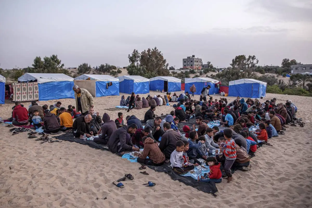 Palestinos deslocados se reúnem para quebrar seu jejum em uma mesa comunitária montada em meio às tendas durante o jantar iftar em massa organizado para palestinos deslocados em Rafah, Gaza, em 27 de março de 2024 [Jehad Alshrafi/Agência Anadolu]
