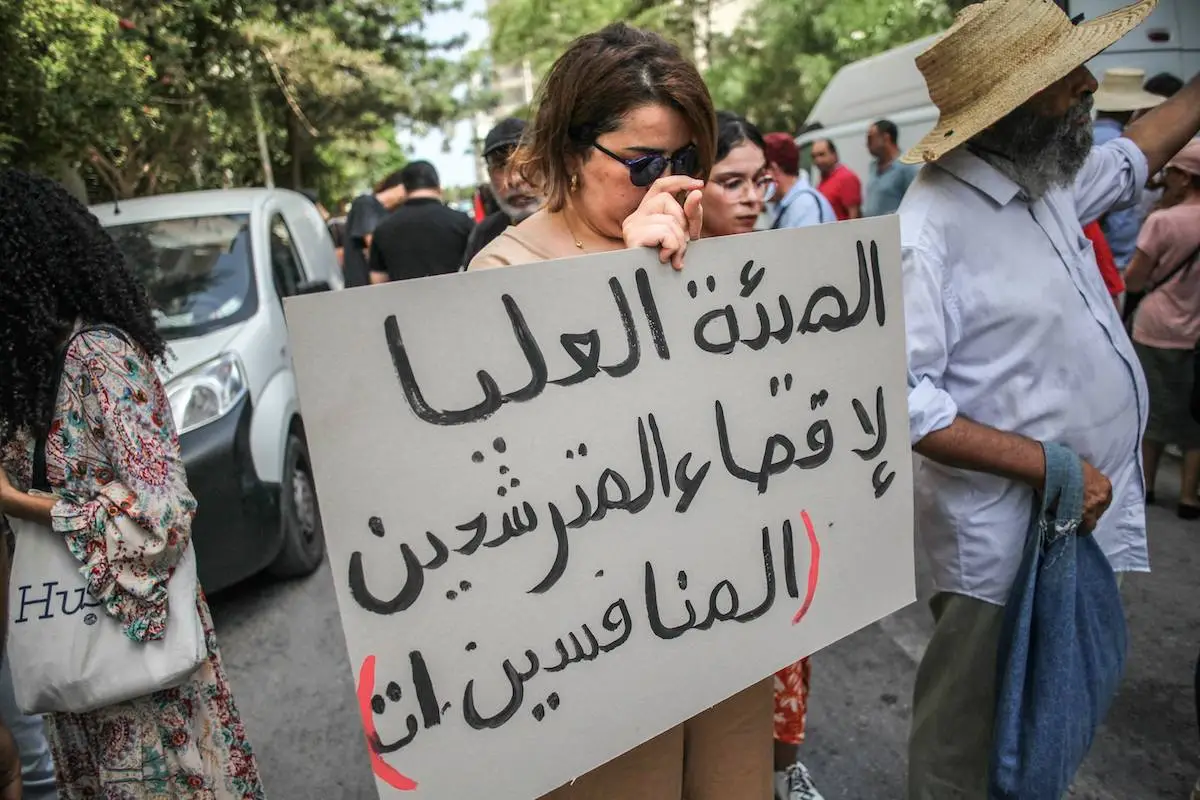 Manifestante segura um cartaz que diz em árabe, ''a Alta Autoridade elimina os candidatos concorrentes'', durante uma manifestação encenada por ativistas de direitos humanos, organizações de direitos das mulheres e grupos da sociedade civil do lado de fora da sede da Alta Autoridade Independente para as Eleições (em francês: ISIE) em Túnis, Tunísia, em 2 de setembro de 2024, para protestar contra a decisão do ISIE de excluir três candidatos às eleições presidenciais e o anúncio da lista final de candidatos oficialmente concorrendo às eleições presidenciais da Tunísia. [ Chedly Ben Ibrahim/NurPhoto via Getty Images]
