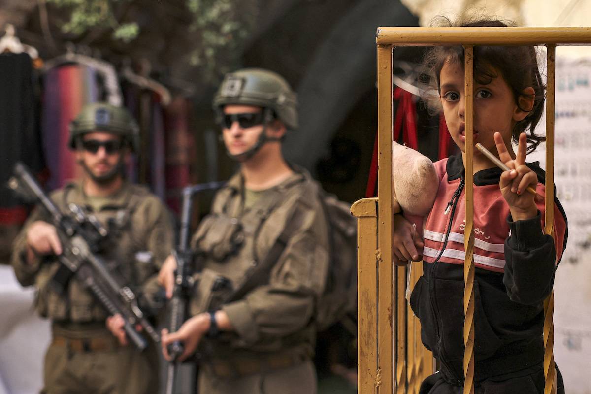 Menina em pé segura uma escova em uma mão e uma boneca de pelúcia na outra em uma sacada perto de soldados israelenses que escoltam colonos israelenses em uma visita guiada ao antigo mercado na parte palestina da cidade velha de Hebron, na Cisjordânia ocupada, em 22 de outubro de 2024, durante o feriado judaico de Sucot, ou a Festa dos Tabernáculos. [Hazem Bader/AFP via Getty Images]
