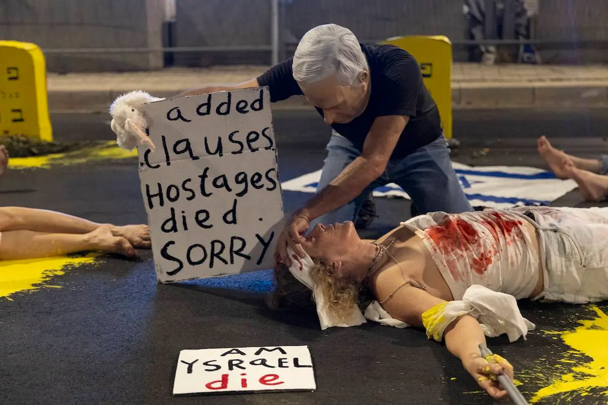 Ativista se deita no chão representando uma refém enquanto um homem se passa pelo primeiro-ministro israelense Benjamin Netanyahu durante uma manifestação pedindo acordo de troca de prisioneiros, em 24 de agosto de 2024 em Tel Aviv, Israel. [Amir Levy/Getty Images]
