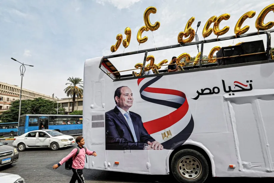 Um estudante caminha perto de um ônibus de campanha eleitoral do presidente egípcio Abdel Fattah al-Sisi, adornado com sua imagem, seu slogan "Viva o Egito" e balões em forma de C, enquanto os apoiadores de Sisi se preparam para um comício em Gizé, cidade gêmea da capital egípcia, em 2 de outubro de 2023. [Foto de Khaled Desouki/AFP via Getty Images]