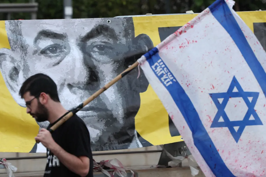 Um manifestante carregando a bandeira israelense passa por um retrato do primeiro-ministro Benjamin Netanyahu durante um protesto antigovernamental em Tel Aviv, em 7 de junho de 2025 [Jack Guez / AFP / Getty Images]