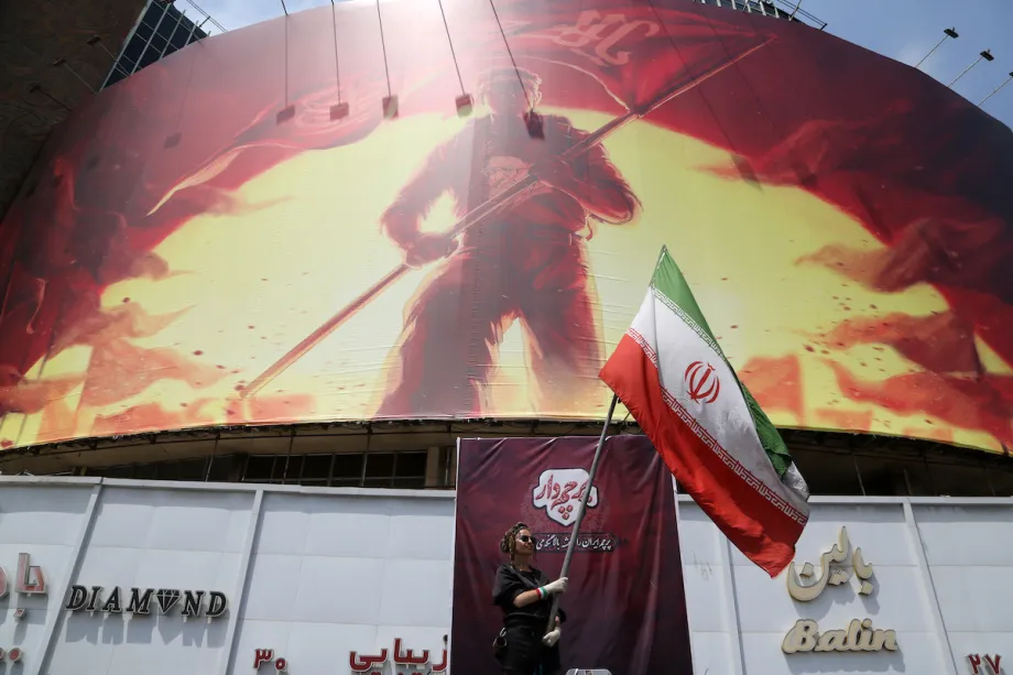 Uma pessoa agita uma bandeira iraniana diante de um enorme painel enquanto a vida cotidiana continua em meio às tensões entre os Estados Unidos e o Irã, em Teerã, Irã, em 6 de agosto de 2026. [Fatemeh Bahrami – Anadolu Agency]