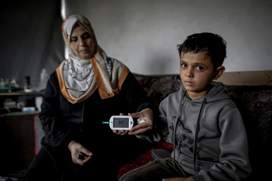 Nihavend, mãe palestina de 39 anos, segura um medidor de glicose no sangue que indica um nível elevado de açúcar, enquanto está sentada ao lado de seu filho Amir, de 10 anos, que tem diabetes tipo 1, na Faixa de Gaza, Palestina, em 26 de outubro de 2026. [Saeed M. M. T. Jaras – Anadolu Agency]